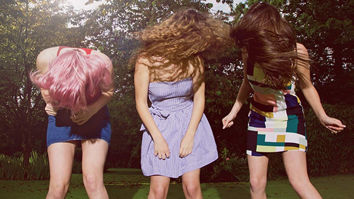 Drei Mädchen schütteln ihre Haare so wild, dass man ihre Gesichter nicht mehr sehen kann. Dabei stehen sie draußen auf einer Wiese