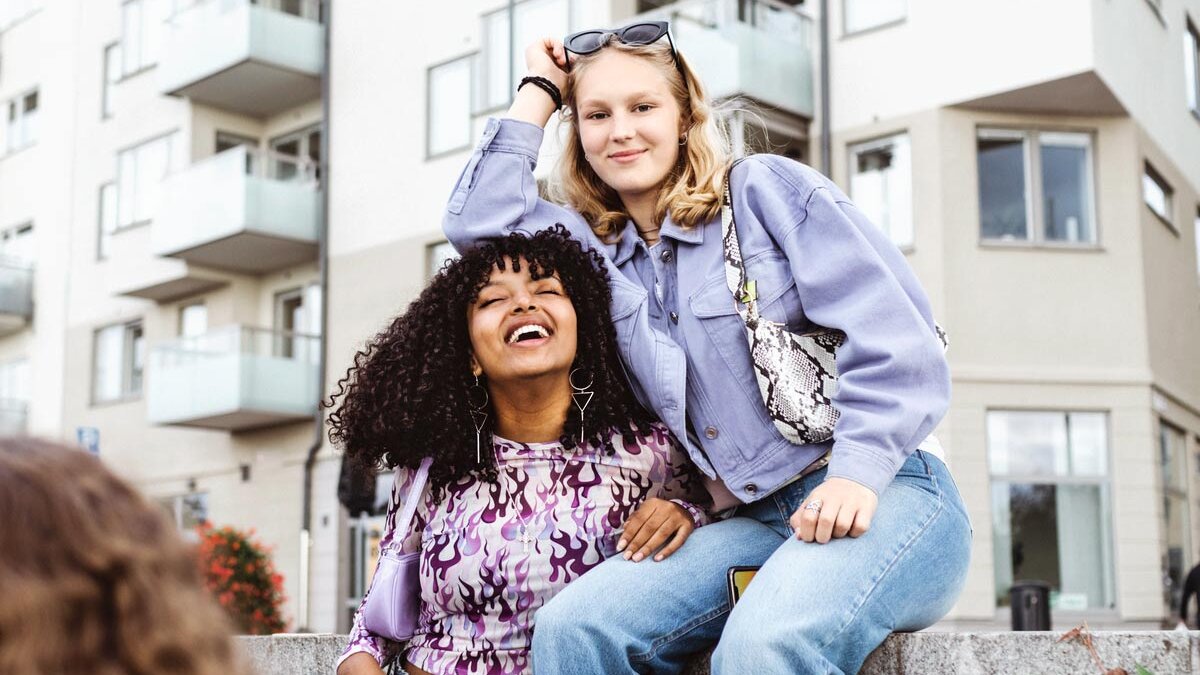 Zwei lächelnde Mädchen sitzen auf einer Mauer. Im Hintergrund sieht man ein Gebäude